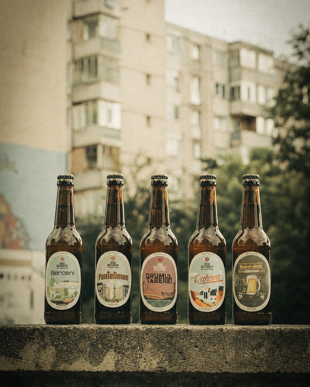 Cinci sticle de Bere București de cartier aliniate pe un parapet, cu un bloc de cărămidă în spate